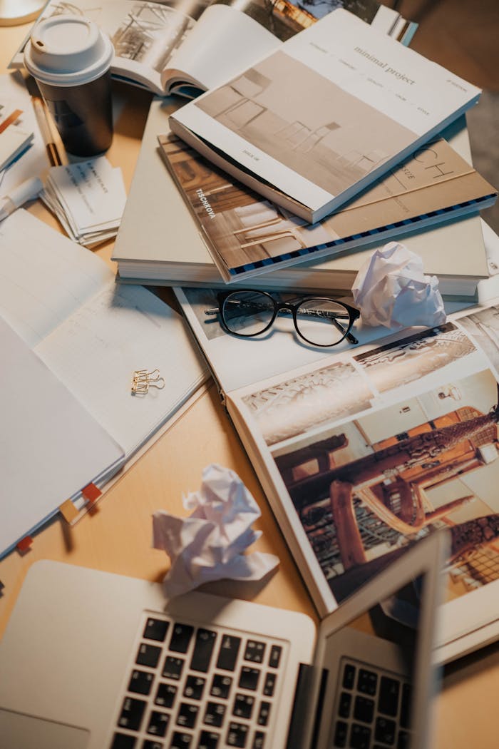 our-story A cluttered desk with books, eyeglasses, crumpled papers, and a laptop, creating an unorganized workspace.